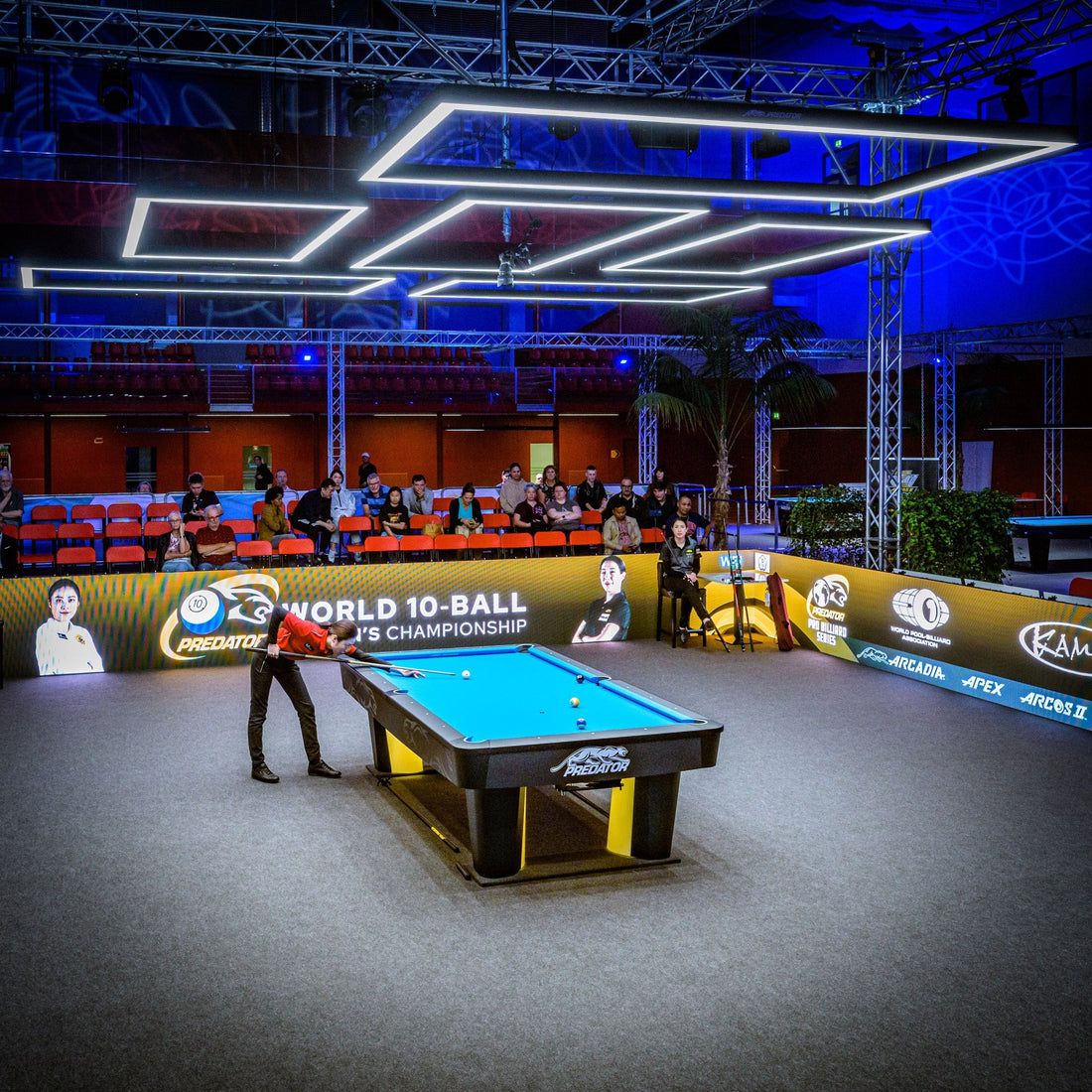 Pool table with a player in a red shirt, surrounded by spectators and event branding.