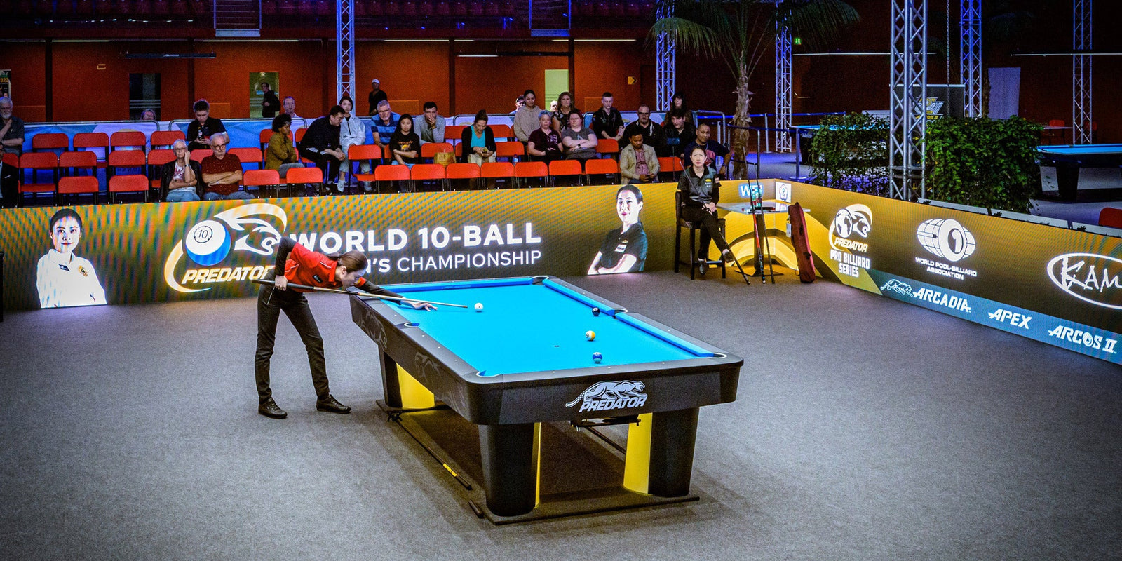 Pool table with a player in a red shirt, surrounded by spectators and event branding.