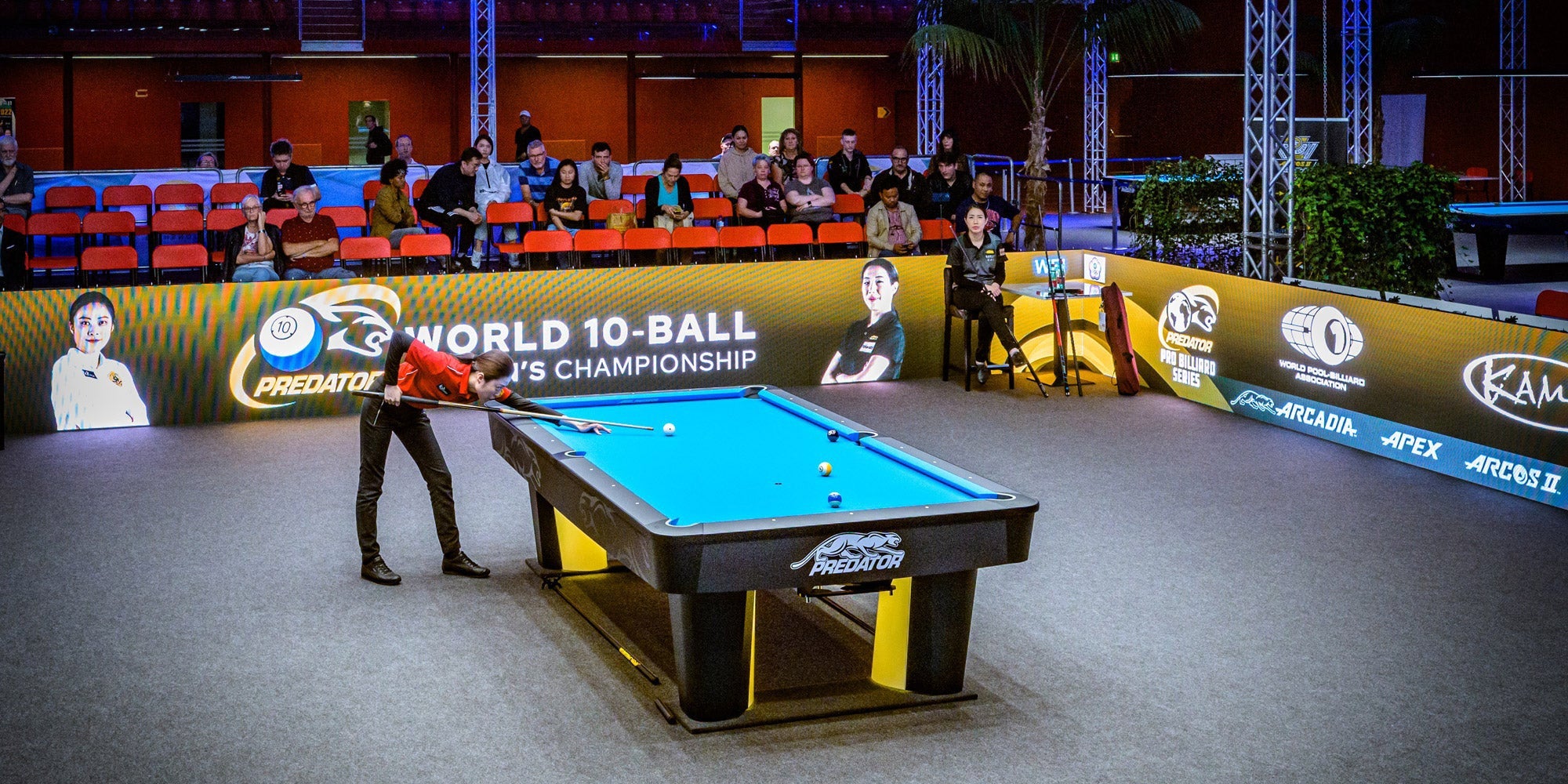 Pool table with a player in a red shirt, surrounded by spectators and event branding.